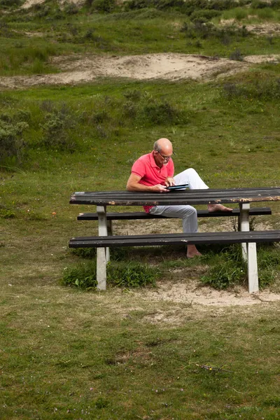 Пенсионер отдыхает и пользуется планшетом за столом в парке — стоковое фото
