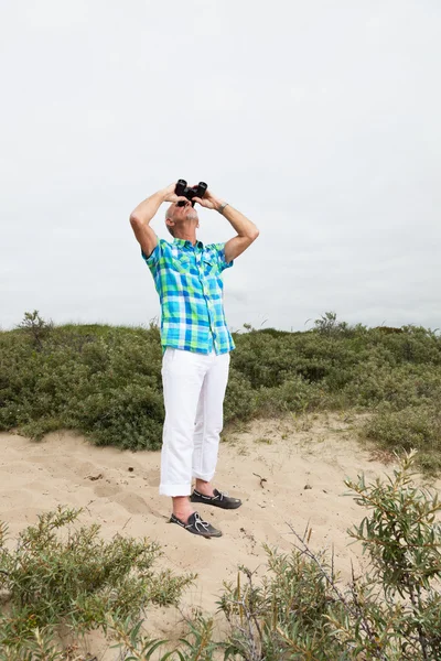 Senior man with beard and glasses using binoculars outdoors in g — Stock Photo, Image