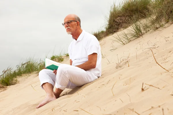 Rentner mit Bart und Brille liest in Grasdüne ein Buch — Stockfoto