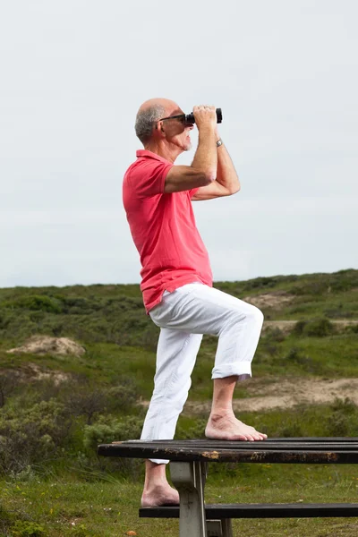 Retired man with beard and glasses using binoculars outdoors in — Stock Photo, Image