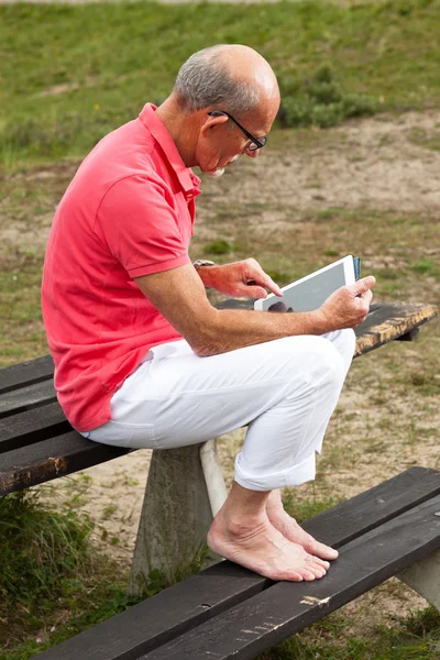 Homem idoso aposentado descansando e usando seu tablet à mesa no parque — Fotografia de Stock