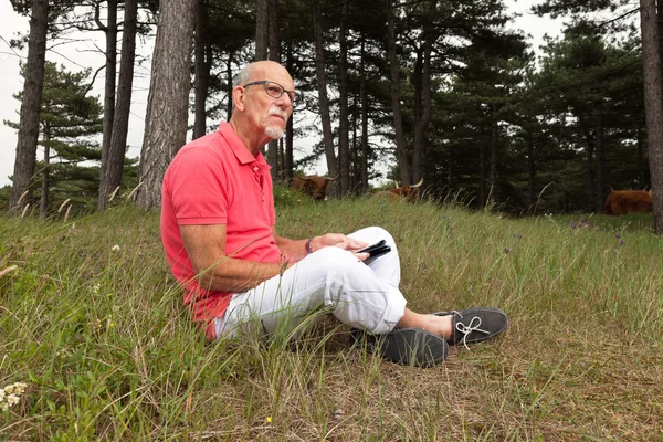 Senior retired man sitting with tablet outdoors in meadow. Scott — Stock Photo, Image