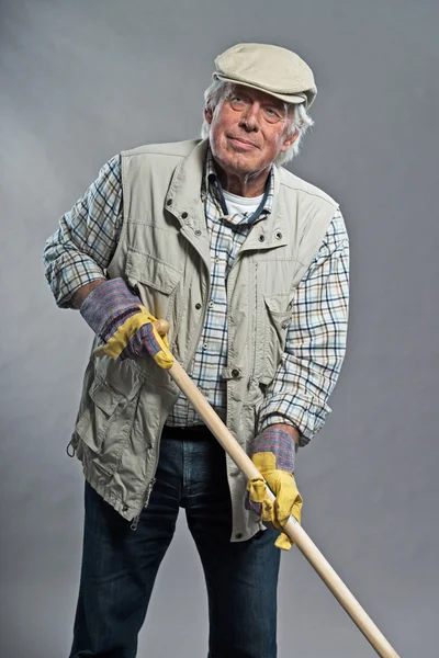 Gardener senior man with hat holding hoe. Studio shot against gr — Stock Photo, Image