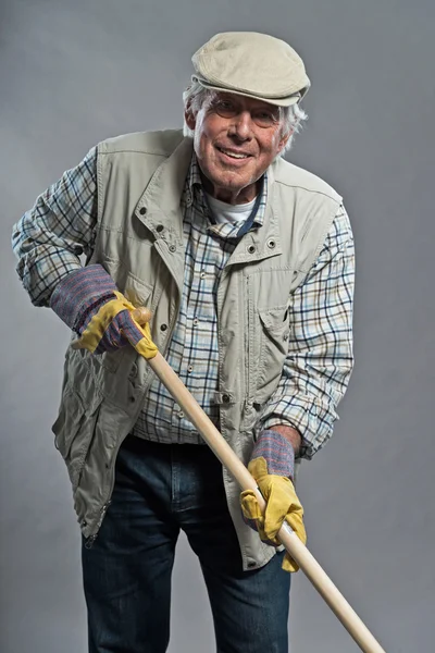 Sonriente jardinero mayor con sombrero sosteniendo azada. Estudio disparo ag — Foto de Stock