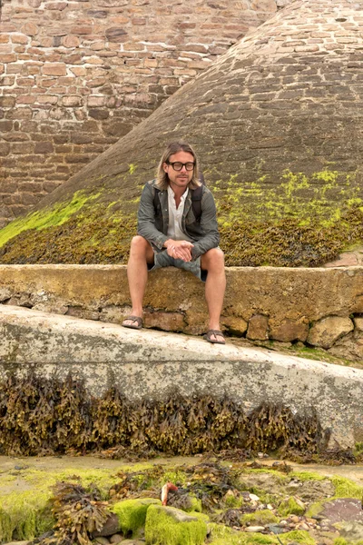 Young man with long blonde hair and glasses sitting on stone wal — Stock Photo, Image