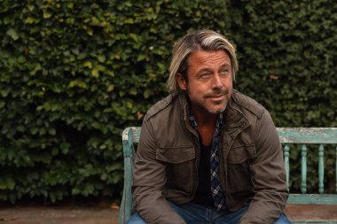 Sarı saçlı, sakallı, yeşil ceketli gülümseyen bir adam yazın arka bahçedeki ahşap bankta oturur..