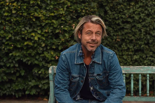 夏には裏庭の木製のベンチに座っているジーンズのジャケットにブロンドの髪とひげのある笑顔の男 — ストック写真