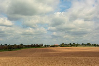 Çayırları, ağaçları ve mavi bulutlu gökyüzünün altında bir evi olan, benekli güneş ışığıyla çıplak bir arazide yuvarlanan bir manzara..