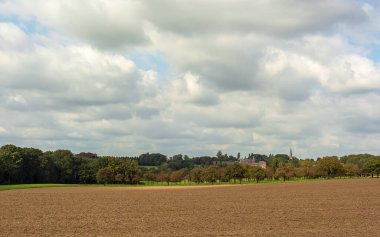 Ağaçları, şatosu ve arka planda bulutlu bir gökyüzünün altında bir kilisesi olan çıplak tarlalar..