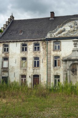 Kasvetli bulutlu gökyüzünün altında yabani otlarla kaplanmış eski, harap ve terk edilmiş bir kır evi..