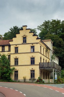 Bulutlu bir gökyüzünün altında, kırsal bir köyde ağaçların olduğu yol boyunca terk edilmiş eski bir ev..