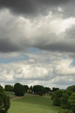 Rolling countryside with meadows, houses and trees under a cloudy sky.
