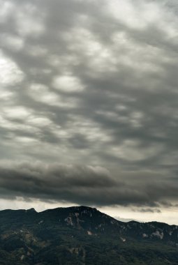 Karanlık bulutlu bir gökyüzünün altında, yamaçta orman olan dağlar..