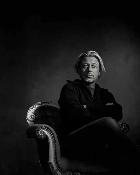 Man with blond hair and a stubble beard in black woolen sweater sits on a vintage bench. Black and white photo.