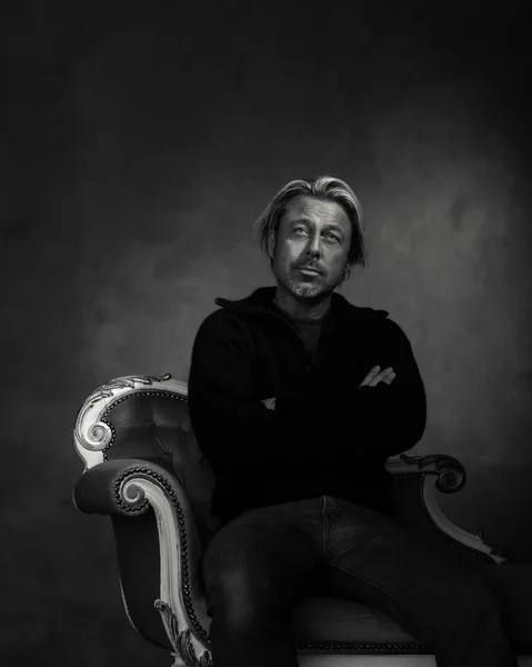 Man with blond hair and a stubble beard in black woolen sweater sits on a vintage bench. Black and white photo.