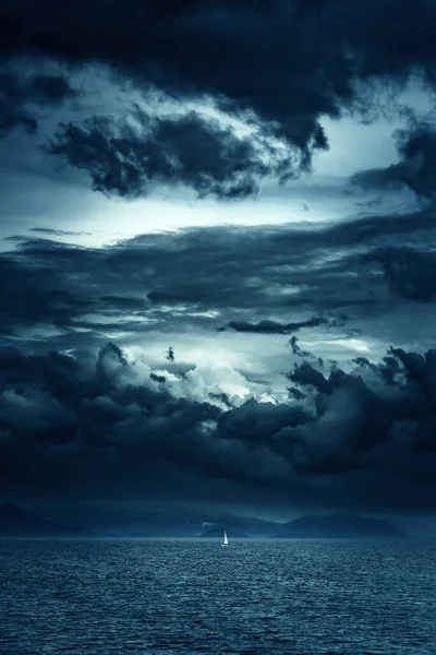 Ocean with a sailboat and mountains on the horizon under a dark cloudy sky.