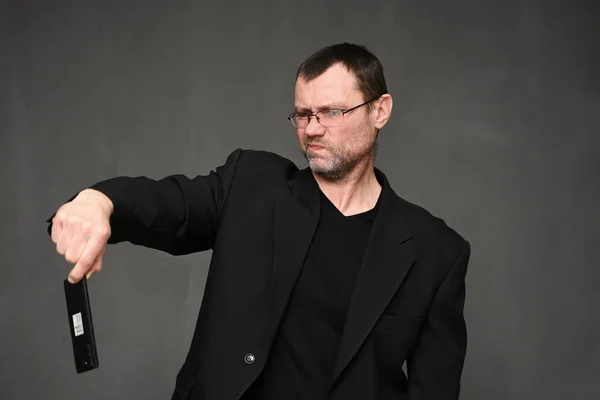 Retrato de un hombre adulto en una chaqueta con gafas con disgusto mira el teléfono en sus manos sobre un fondo gris — Foto de Stock
