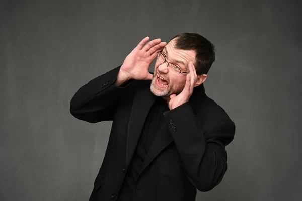 Blanke man in een zwart jasje met bril schreeuwend naar de zijkant met emoties op een grijze achtergrond — Stockfoto