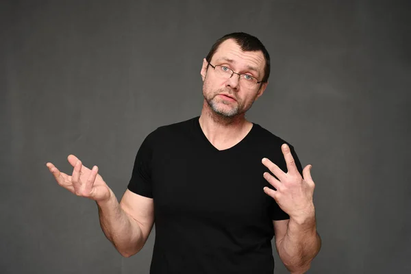 Blanke man in een zwart t-shirt met bril in gesprek met emoties op een grijze achtergrond — Stockfoto