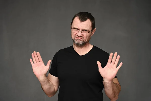 Volwassen man in een zwart t-shirt met bril shows stop met zijn handen op een grijze achtergrond — Stockfoto