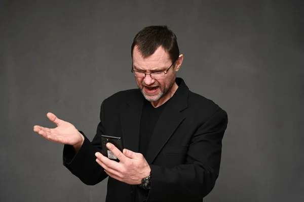 Caucasian man in a black jacket with glasses looks displeased at the phone with emotions on a gray background Stock Image