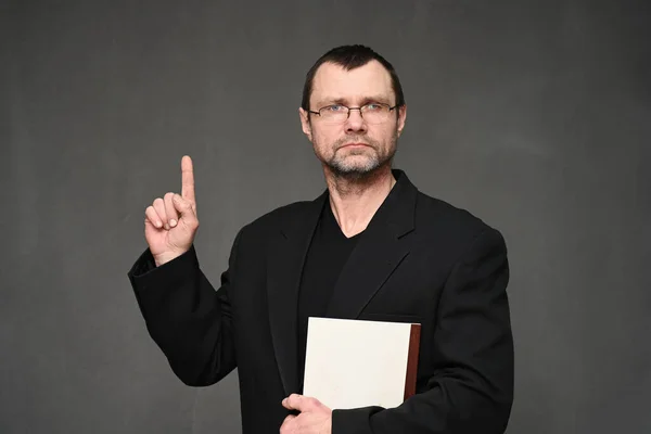 Een volwassen man in een jasje kijkt naar de camera met een map in zijn handen, wijzend met zijn vinger omhoog. Portret op een grijze achtergrond in de studio — Stockfoto