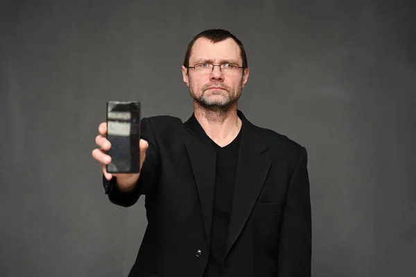 Hombre de negocios caucásico en una chaqueta mira a la cámara y muestra la pantalla de un teléfono inteligente a la cámara. Retrato de un hombre adulto sobre un fondo gris en el estudio — Foto de Stock