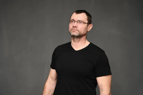 Un hombre adulto con gafas mira hacia otro lado. Retrato de un hombre caucásico con una camiseta negra sobre un fondo gris en el estudio —  Fotos de Stock