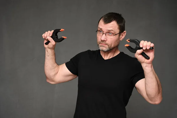 Een volwassen man met een bril en een zwart t-shirt toont draadscharen op een grijze achtergrond. — Stockfoto