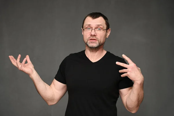 Un hombre adulto en gafas con emociones habla con la cámara. Retrato de un hombre caucásico sobre un fondo gris en el estudio Imágenes de stock libres de derechos