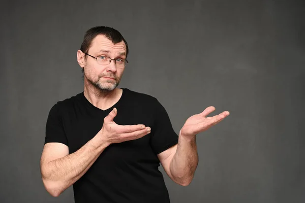 Дорослий чоловік в окулярах показує з емоціями руки в бік. Портрет кавказького чоловіка в чорній футболці на сірому фоні в студії Стокове Зображення