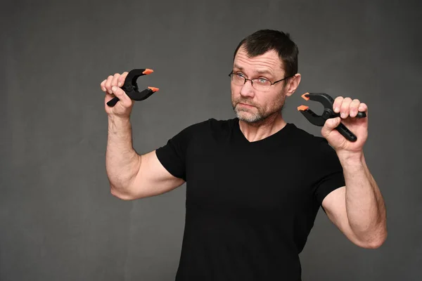 An adult man in glasses and a black t-shirt shows wire cutters on a gray background. Royalty Free Stock Photos