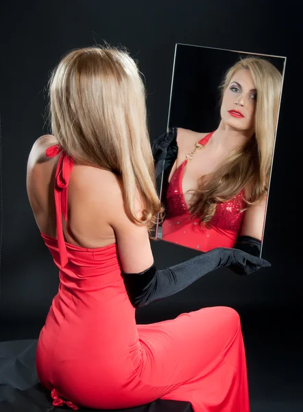 Beauiful young woman in red dress looks into the mirror on dark background — Stock Photo, Image