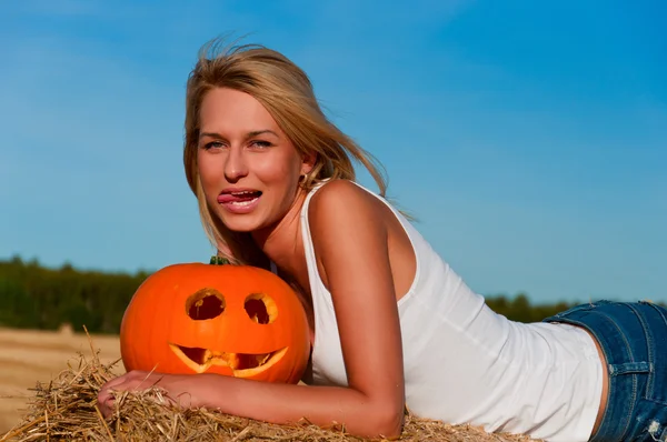 Woman in jeans shorts posing on a bale with pumpkin — Stock Photo, Image