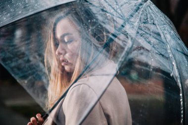 Paltolu genç, narin, güzel bir kız parkta şeffaf bir şemsiyeyle yağmurda yürüyor.