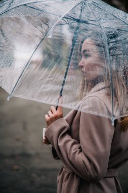 Paltolu genç, narin, güzel bir kız parkta şeffaf bir şemsiyeyle yağmurda yürüyor.