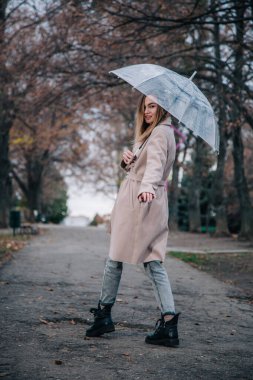 Paltolu genç, narin, güzel bir kız parkta şeffaf bir şemsiyeyle yağmurda yürüyor.
