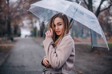 Paltolu genç, narin, güzel bir kız parkta şeffaf bir şemsiyeyle yağmurda yürüyor.