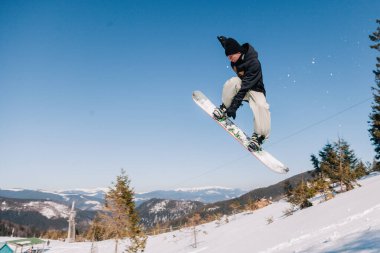 Siyah ceketli bir adam, bir snowboard 'a atlayıp, dağların arka planına doğru atlıyor.