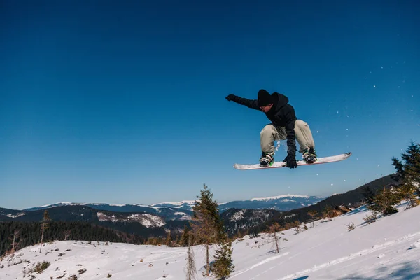 Ein Mann in schwarzer Jacke springt auf ein Snowboard vor der Kulisse der Berge und hält einen vorderen Griff — Stockfoto