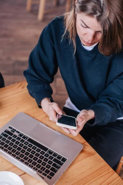 Młoda piękna dziewczyna w czarnym swetrze w kawiarni pracuje z laptopem i telefonem — Zdjęcie stockowe