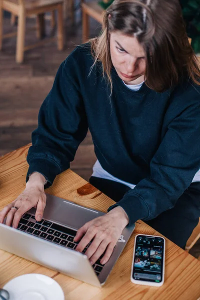 Ένα νεαρό όμορφο κορίτσι σε ένα μαύρο πουλόβερ σε ένα καφέ δουλεύει με ένα φορητό υπολογιστή και ένα τηλέφωνο — Φωτογραφία Αρχείου