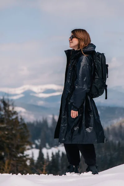 Una joven con gafas de sol y una chaqueta negra en una estación de esquí con el telón de fondo de las montañas — Foto de Stock