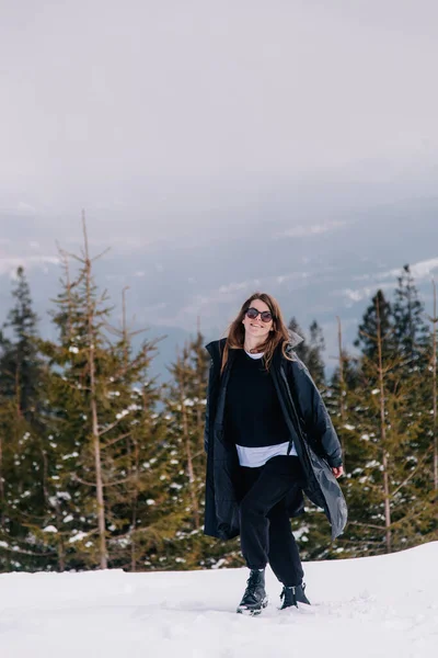 Una joven con gafas de sol y una chaqueta negra en una estación de esquí con el telón de fondo de las montañas — Foto de Stock