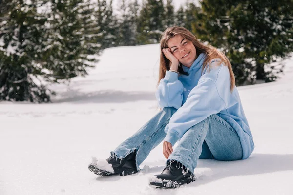 A young, beautiful girl in the mountains sits in the snow among the trees — Stock Photo, Image