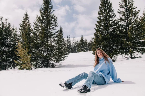 A young, beautiful girl in the mountains sits in the snow among the trees — Stock Photo, Image