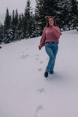 Genç, ince, uzun saçlı bir kız dağlardaki bir kış ormanında karda koşuyor..