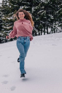 Genç, ince, uzun saçlı bir kız dağlardaki bir kış ormanında karda koşuyor..