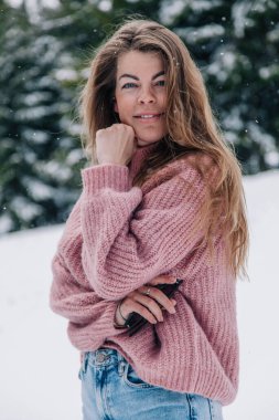 Genç, güzel, uzun saçlı pembe kazaklı bir kız dağlardaki bir kış ormanında poz veriyor.