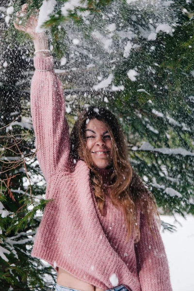 Ein junges, fröhliches Mädchen, das unter den Ästen einer Fichte in den Bergen mit Schnee bestreut wird — Stockfoto